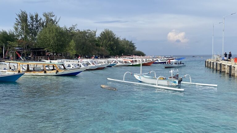 Boats docked in clear turquoise water at Gili Trawangan, with visible coral reef beneath the surface near the harbor